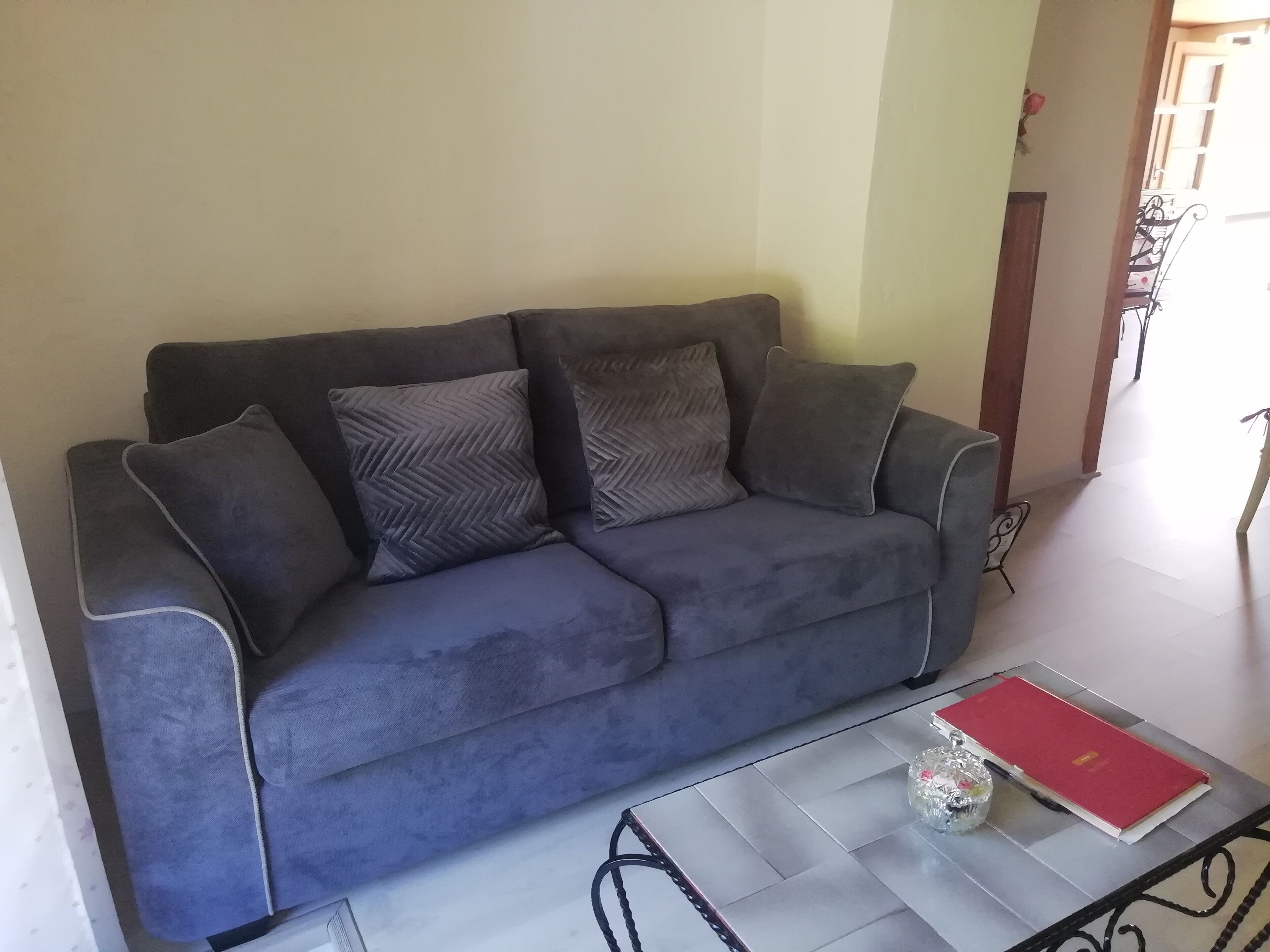 A grey sofa with patterned cushions in a room with a tiled floor and a small table with a red book and a glass object.