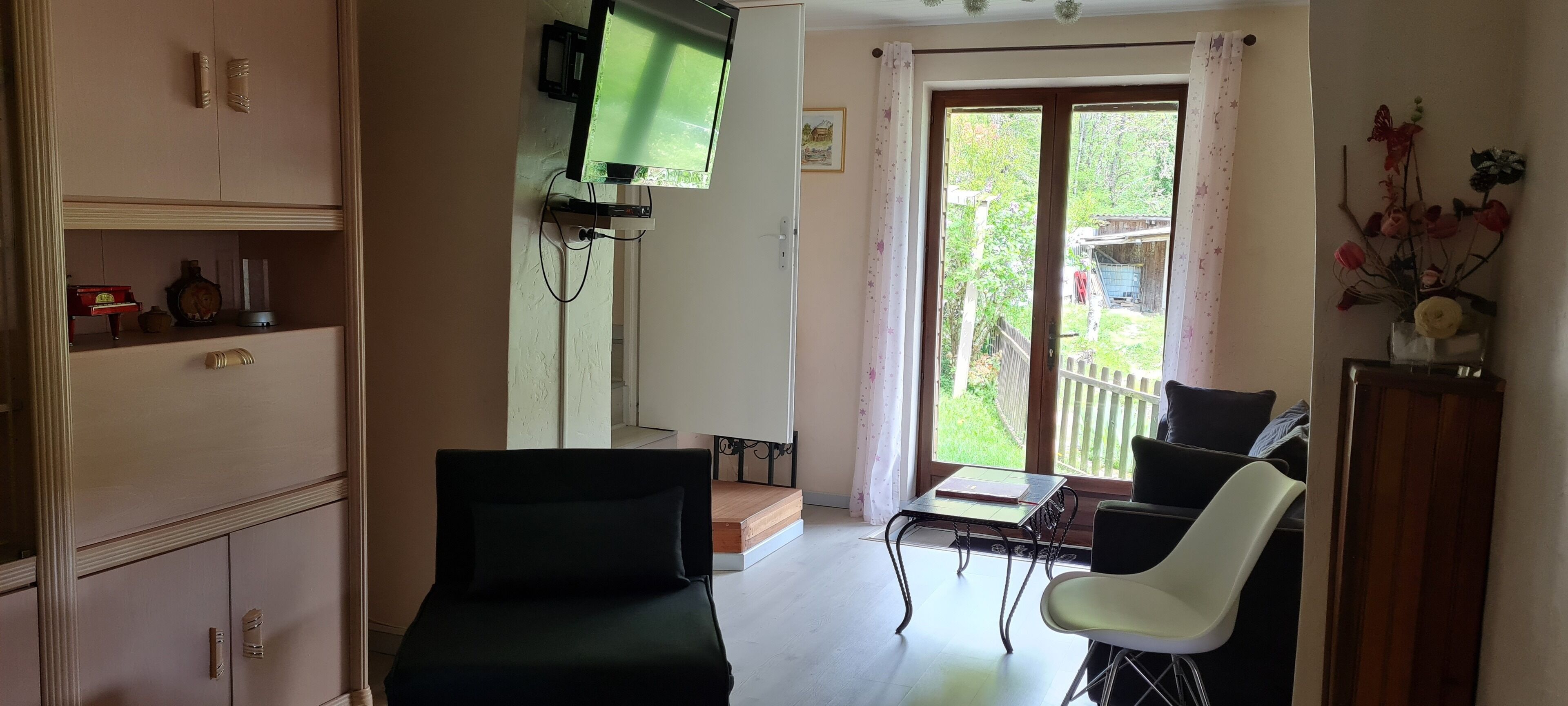 A room with a TV mounted on the wall, a table with chairs, a cabinet with decorative items, and a view of the outside through a glass door.