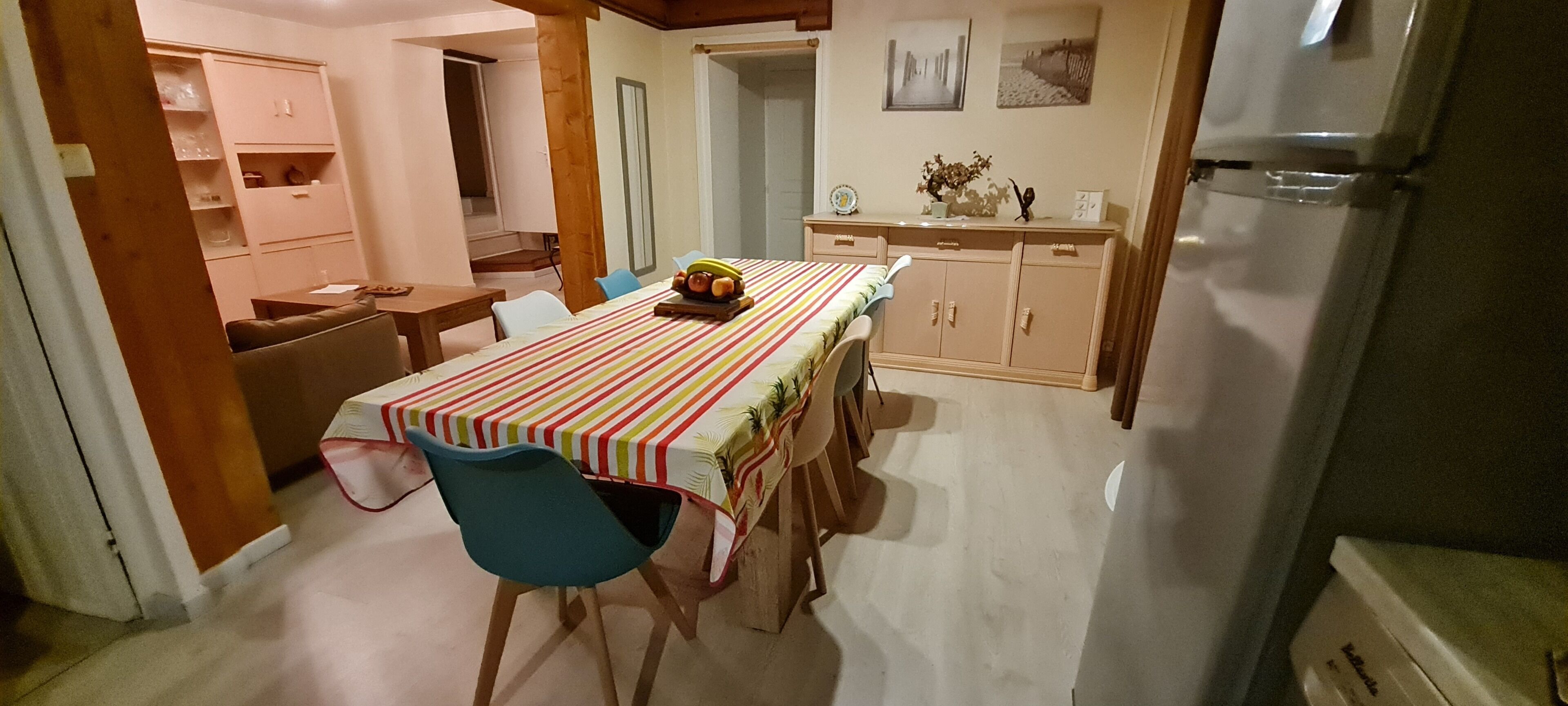 Ein Essbereich mit gestreiftem Tischleintuch, Holzsesseln, einem Schrank mit Dekoration und einem Kühlschrank.