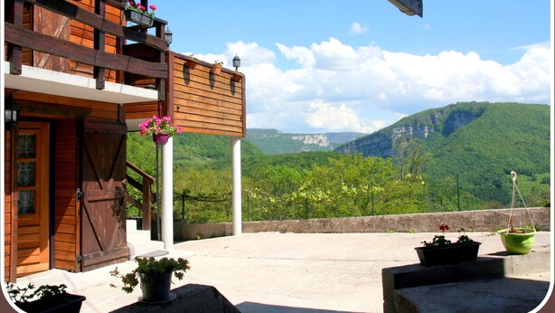 A wooden building with balconies, a concrete terrace, and a view of green hills and mountains.