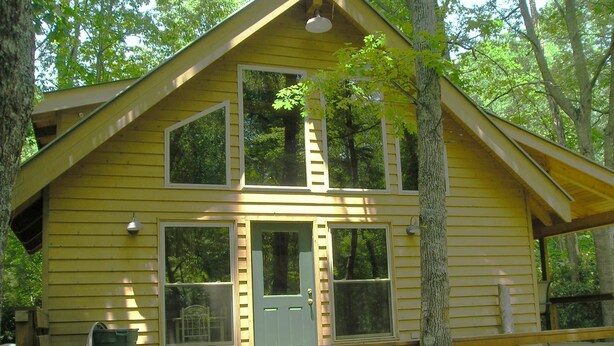 Une cabane jaune avec un porche à l’avant, de grandes fenêtres et une porte.