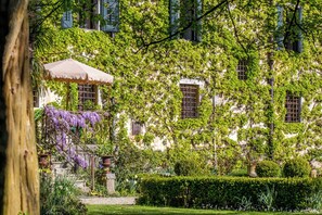A building with ivy-covered walls, a garden with a hedge, and a pergola with a canopy.