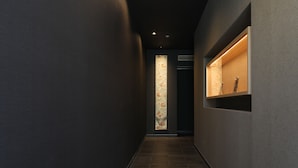 A narrow hallway with dark walls and a tiled floor. There is a vertical frosted glass panel with a floral pattern on the left and a shelf with a sculpture on the right. - Hotel Cocon (Fukuoka)