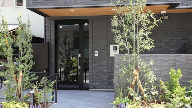 A modern building entrance with a dark facade, a wooden overhang, and a landscaped garden area with various plants and a small tree. - Hotel Cocon (Fukuoka)