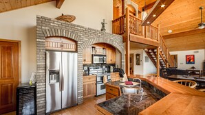 A spacious kitchen with a stone fireplace, wooden cabinets, and a stainless steel refrigerator.