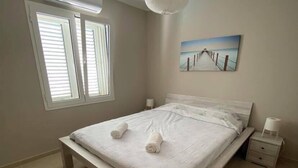 Quarto com cama, janela com persianas, ar condicionado, quadro pendurado na parede e mesa de cabeceira com luminária.