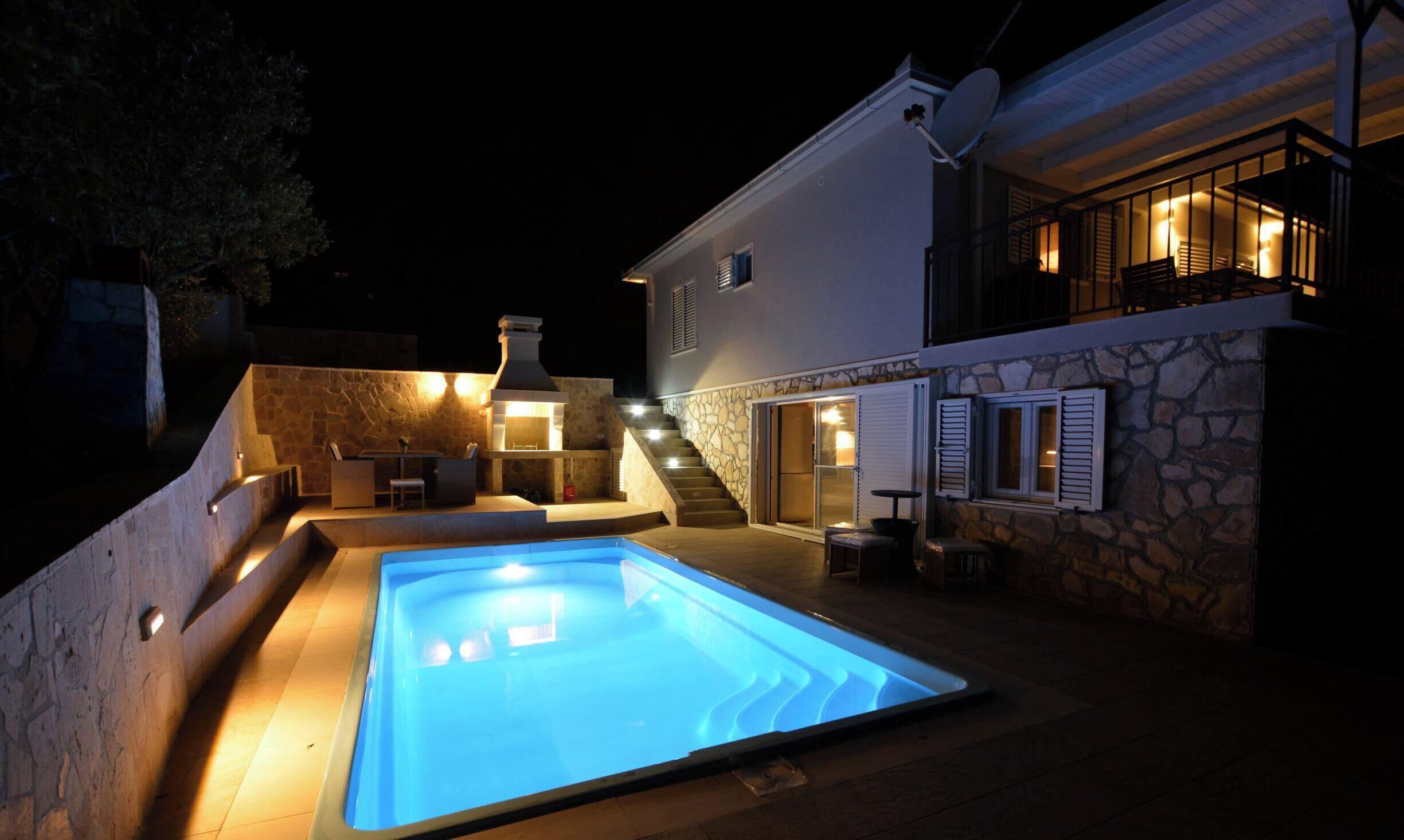 A swimming pool with a stone wall, a barbecue area, and a staircase leading to a second-floor balcony.