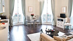 A spacious living room with a dining area, large windows with curtains, a chandelier, and a television. - Perfectly Parisian, Bright & Elegant with Balcony (Paris)