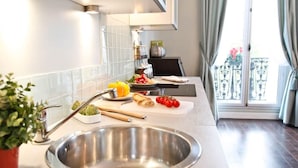 A modern kitchen with a stainless steel sink, a countertop with fresh vegetables, and a view of a balcony with a railing. - Perfectly Parisian, Bright & Elegant with Balcony (Paris)