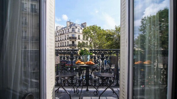 A balcony with a table and chairs, a potted plant, and a view of buildings. - Perfectly Parisian, Bright & Elegant with Balcony (Paris)