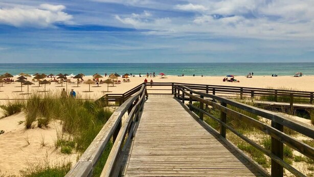 A wooden boardwalk leading to a beach with umbrellas and people.