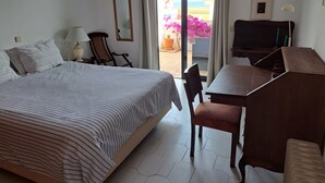 A hotel room with a bed, a desk, a chair, a TV, and a balcony with a view of the sea.