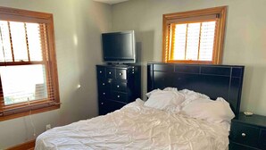Une chambre à coucher comprenant un lit, une commode, un téléviseur et deux fenêtres équipées de stores.