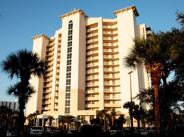 A multi-story hotel with a central tower, surrounded by palm trees and a parking area.