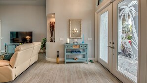 Ein Wohnzimmer mit einem beigen Sofa, einem Fernseher, einem blauen Sideboard und einem großen Fenster mit Blick auf Palmen.