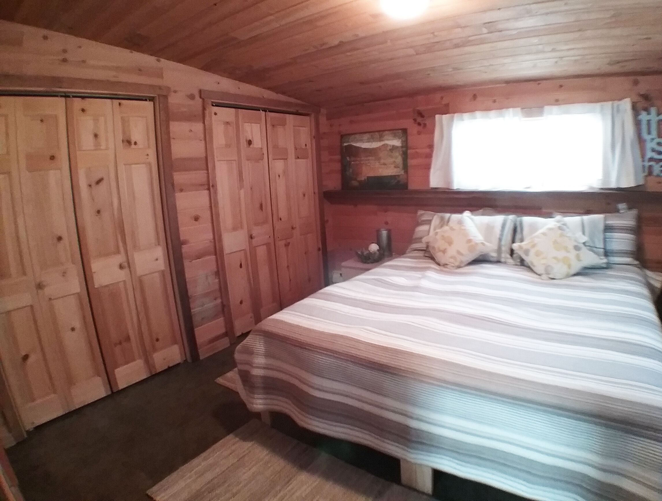 A wooden cabin bedroom with a bed, pillows, a nightstand, and a window with curtains.