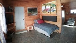 A cozy cabin room with a bed, a small table, a chair, a door, and a painting on the wall.