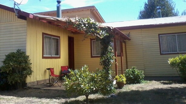 Una casa amarilla de un piso con una antena parabólica, un pequeño jardín con un árbol y macetas, y una silla plegable roja en el exterior.