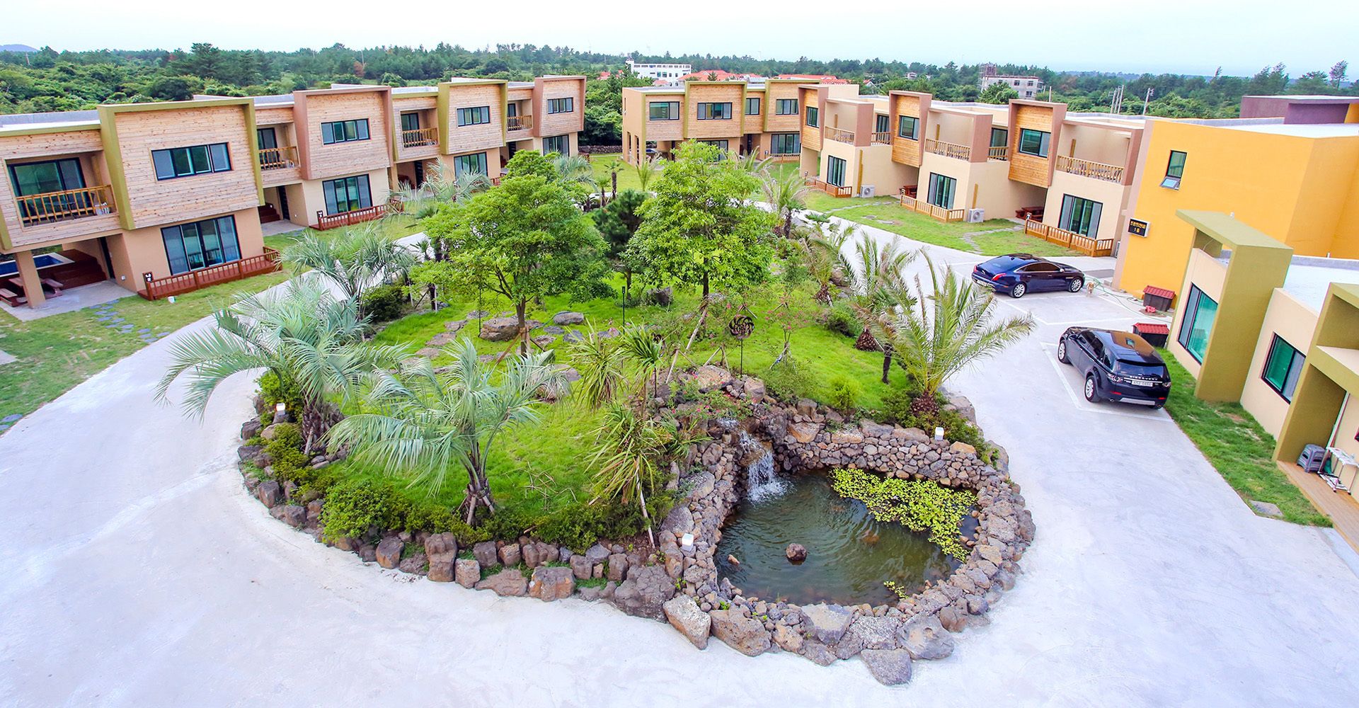 A residential complex with multiple two-story buildings, a central landscaped area with a small pond, and a parking area with cars.