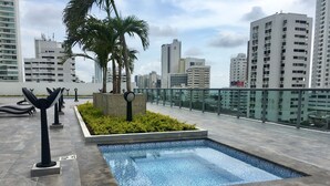 Piscina no terraço com piso ladrilhado, cercada por vegetação e palmeiras, com um horizonte urbano ao fundo.