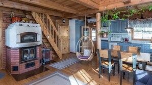 A wooden cabin interior with a brick fireplace, wooden dining table, hanging chair, and a kitchen area.