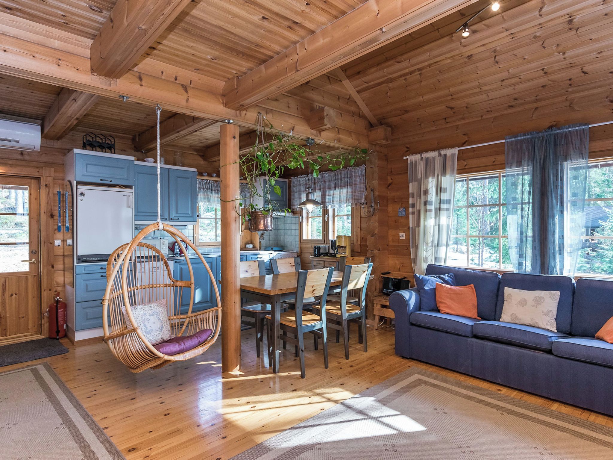 A wooden cabin interior with a dining area, a hanging chair, a sofa, and a kitchen.
