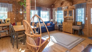 A cozy wooden cabin interior with a dining area, a hanging chair, a sofa, and a kitchen area.