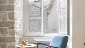 Een kamer met een stenen muur, een houten vloer, een blauwe stoel, een klein tafeltje met sinaasappels en een raam met uitzicht op gebouwen.
