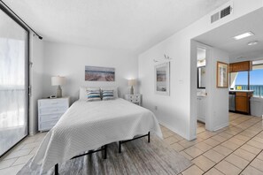 A bedroom with a bed, bedside tables, a lamp, a painting, and a view of the kitchen.