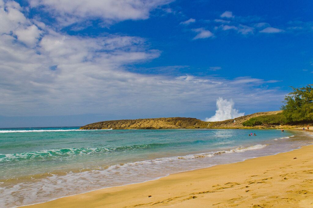 Une plage à l’eau turquoise et limpide, un rivage sablonneux et, au loin, un promontoire rocheux.