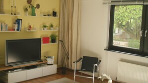 A living room with a flat-screen TV on a wooden stand, a black chair, a small table with a vase of flowers, and a window with a view of greenery. - Living comfortably in Oberwalluf, enjoying the Rheingau, experiencing the Rhine - Main! (Walluf)