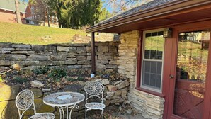Ein Steinhaus mit einer roten Tür, einer kleinen Terrasse mit zwei Metallstühlen und einem Tisch sowie einer Steinmauer.