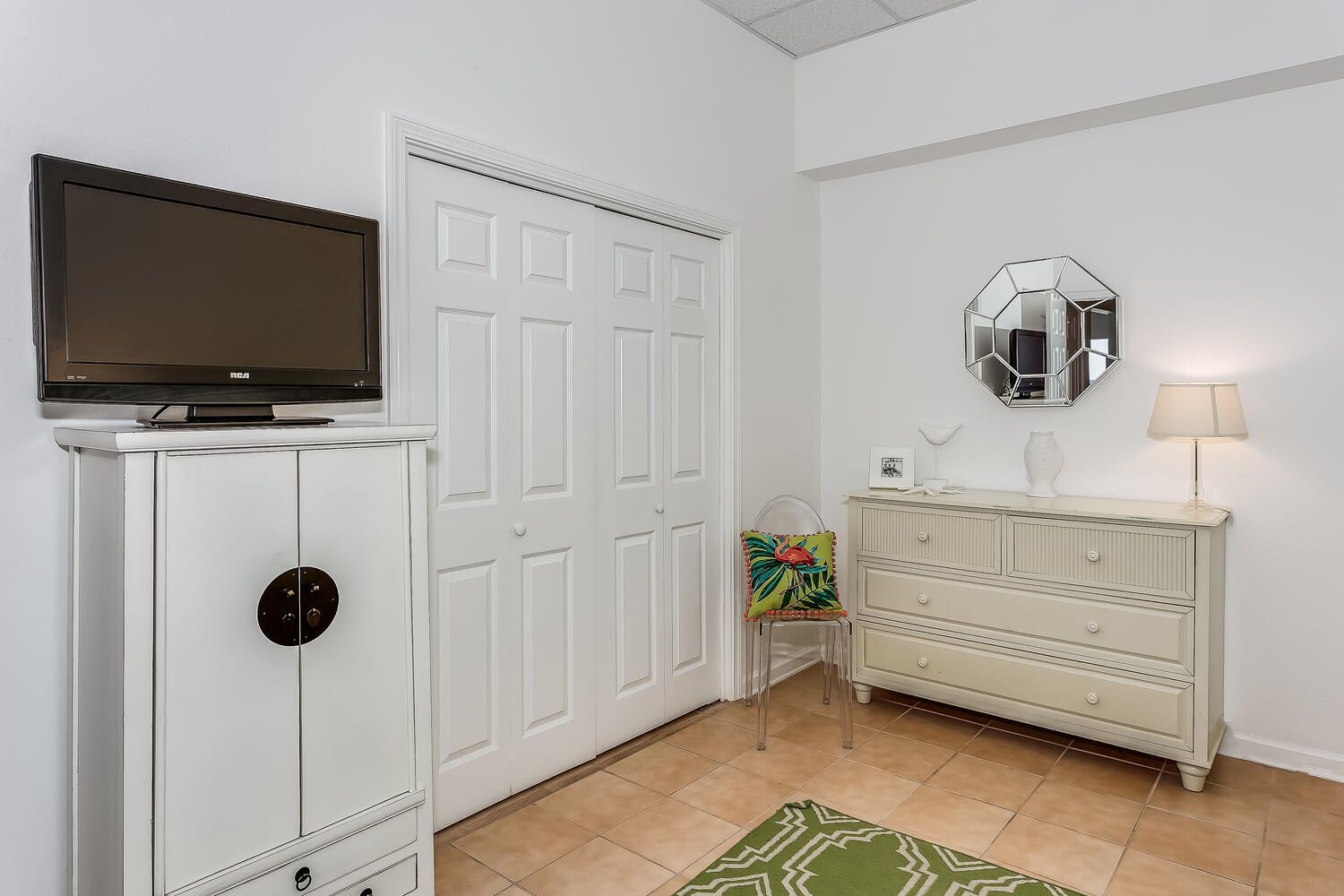 A room with a white wardrobe, a television on top, a mirror above a dresser, and a small table with a lamp.