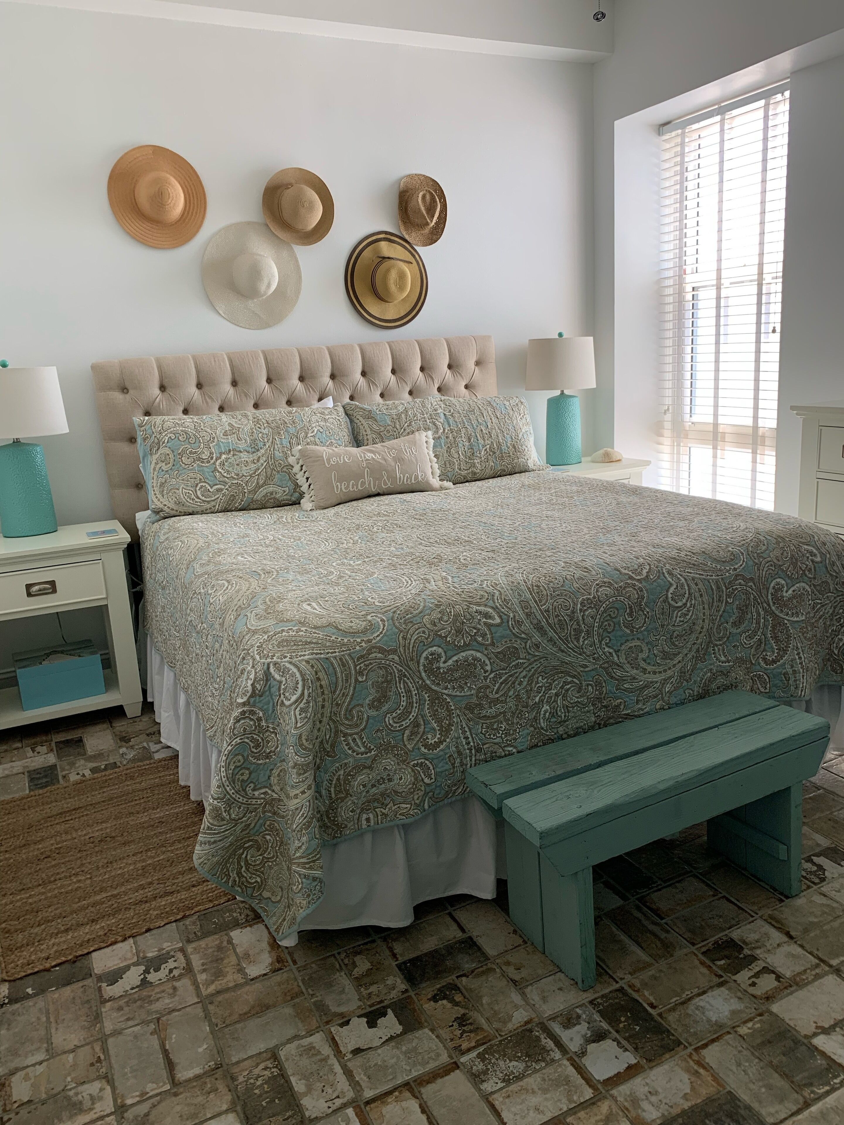 Una camera da letto con una testiera trapuntata, un letto con biancheria a motivi, una panca e cappelli appesi al muro.