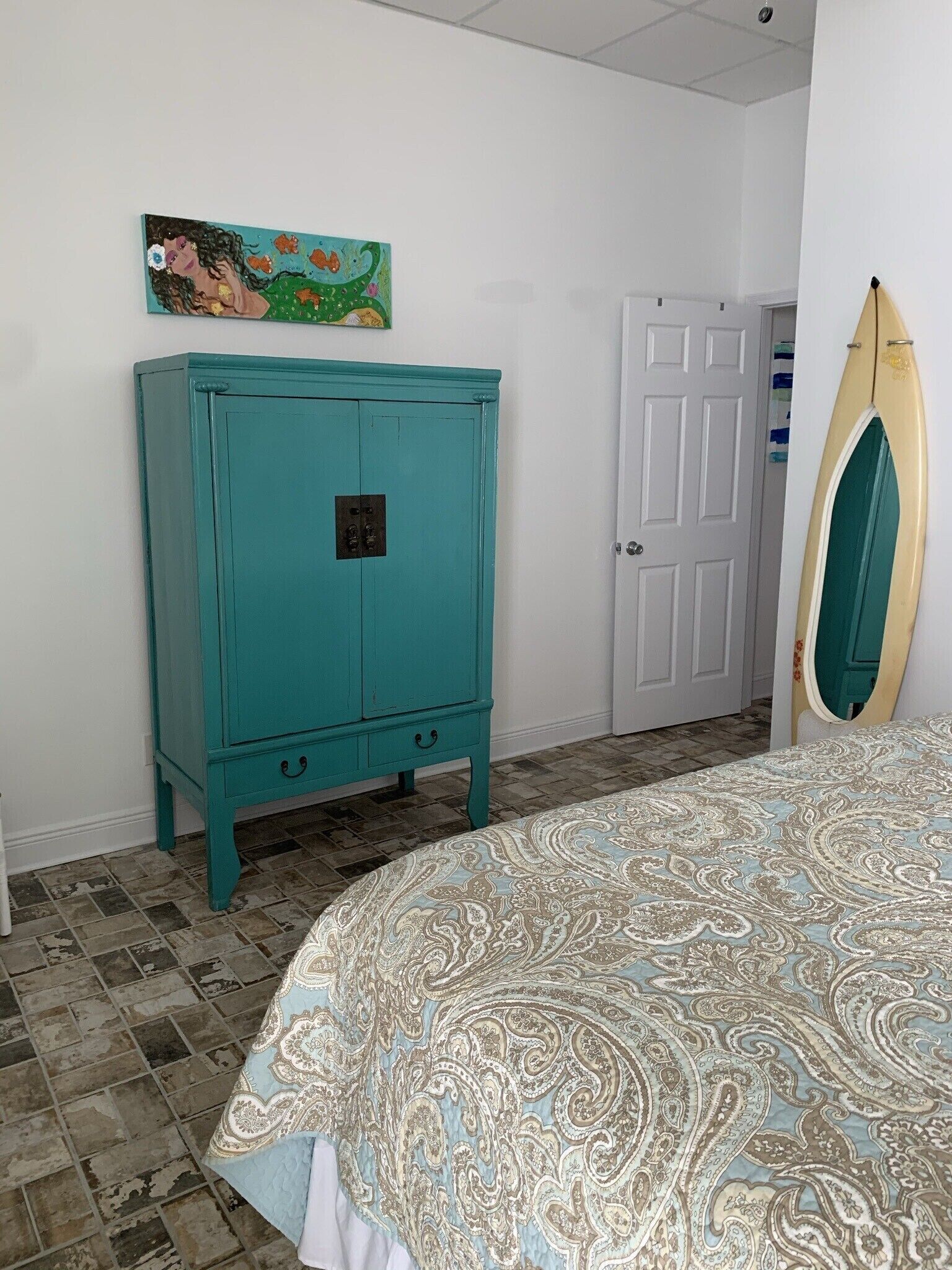 Ein Schlafzimmer mit einem türkisblauen Schrank, einem Bett mit gemustertem Überwurf, einer weißen Tür und einem Wandbild.