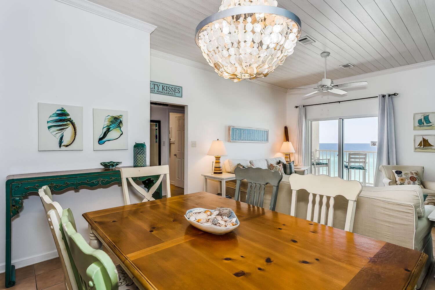 Un comedor con mesa de madera, sillas y una lámpara de araña. Se observan obras de arte en las paredes y un letrero que indica la ubicación.