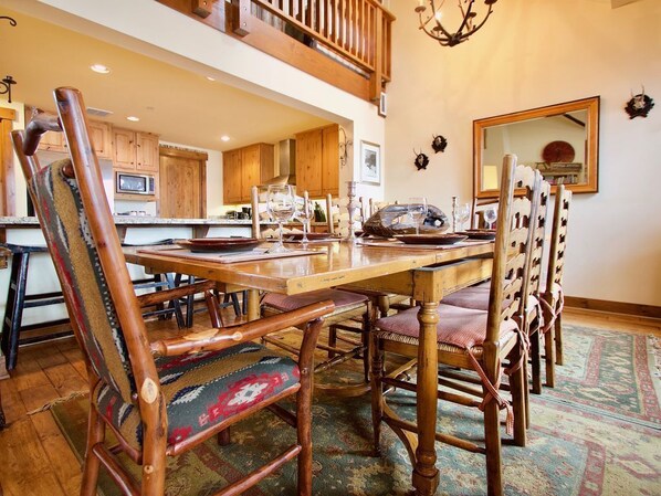 A dining area with wooden chairs and a table set for a meal, a kitchen in the background, and a chandelier.