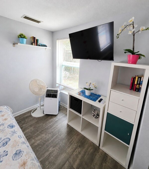 Une chambre à coucher avec un lit, un bureau avec une télévision, un ventilateur et des étagères remplies de livres et de plantes.