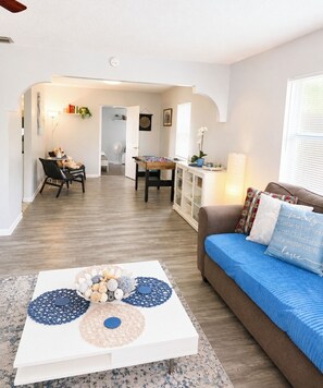 A living room with a brown sofa, a white coffee table with decorative items, a dining area with a table and chairs, and a kitchen area with cabinets and a countertop.