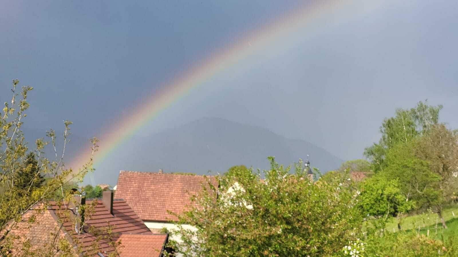 Une montagne sur laquelle se dessine un arc-en-ciel, entourée de verdure et de bâtiments aux toits recouverts de tuiles rouges.