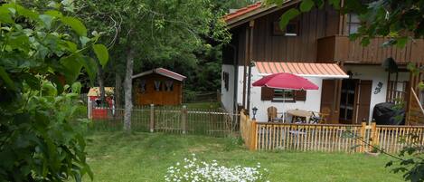 Une maison en bois avec un toit rouge, une cour clôturée et un petit jardin agrémenté de fleurs blanches.