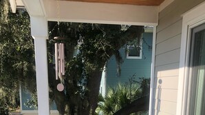 Un divano in vimini con cuscini su un pontile in legno, un campanello eolico sospeso e vista su un edificio verde e alberi.