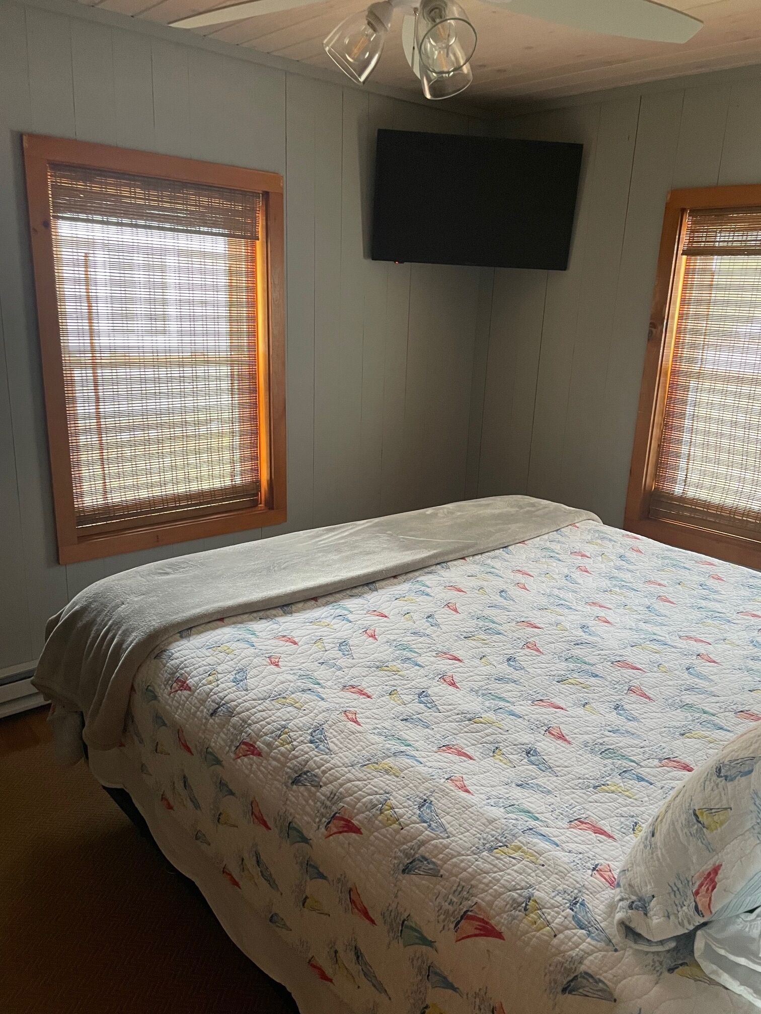 Una camera da letto con un letto, due finestre con tende, una ventola a soffitto e un televisore a schermo piatto montato a parete.