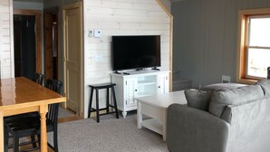 Un salon avec une table à manger en bois, un canapé gris et un téléviseur à écran plat posé sur un meuble blanc.