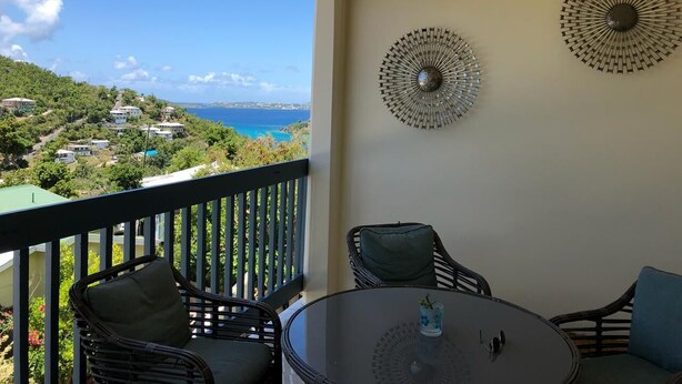 Un balcon agrémenté de fauteuils et d’une table en osier, offrant une vue sur un paysage côtier avec des maisons et la mer.