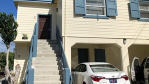 Un bâtiment à deux étages, avec une façade de couleur beige et des bordures bleues. Un escalier mène à l’entrée, et une voiture blanche est garée dans l’allée.