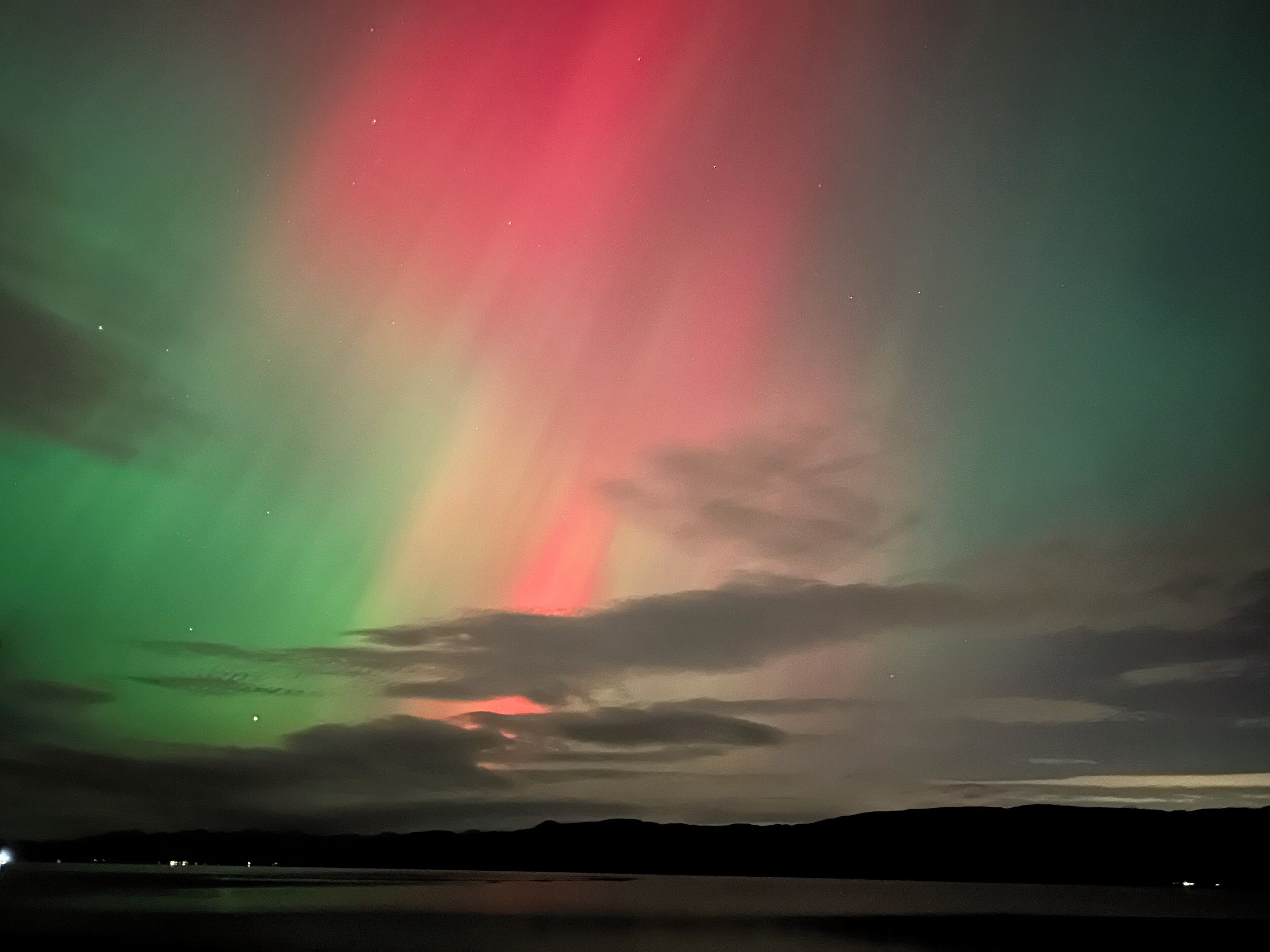 A night sky with the aurora borealis over a calm body of water and distant mountains.