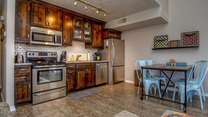 A compact kitchen with wooden cabinets, stainless steel appliances, a dining area with blue chairs, and a chessboard on the wall. - Athlete, Family, and Pet Friendly - Something for everyone! (St. George)