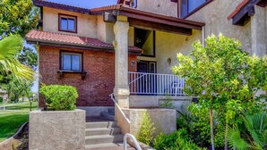 A two-story building with a brick facade, a red-tiled roof, and a small porch with a white railing. There are steps leading up to the entrance, and the building is surrounded by greenery. - Athlete, Family, and Pet Friendly - Something for everyone! (St. George)