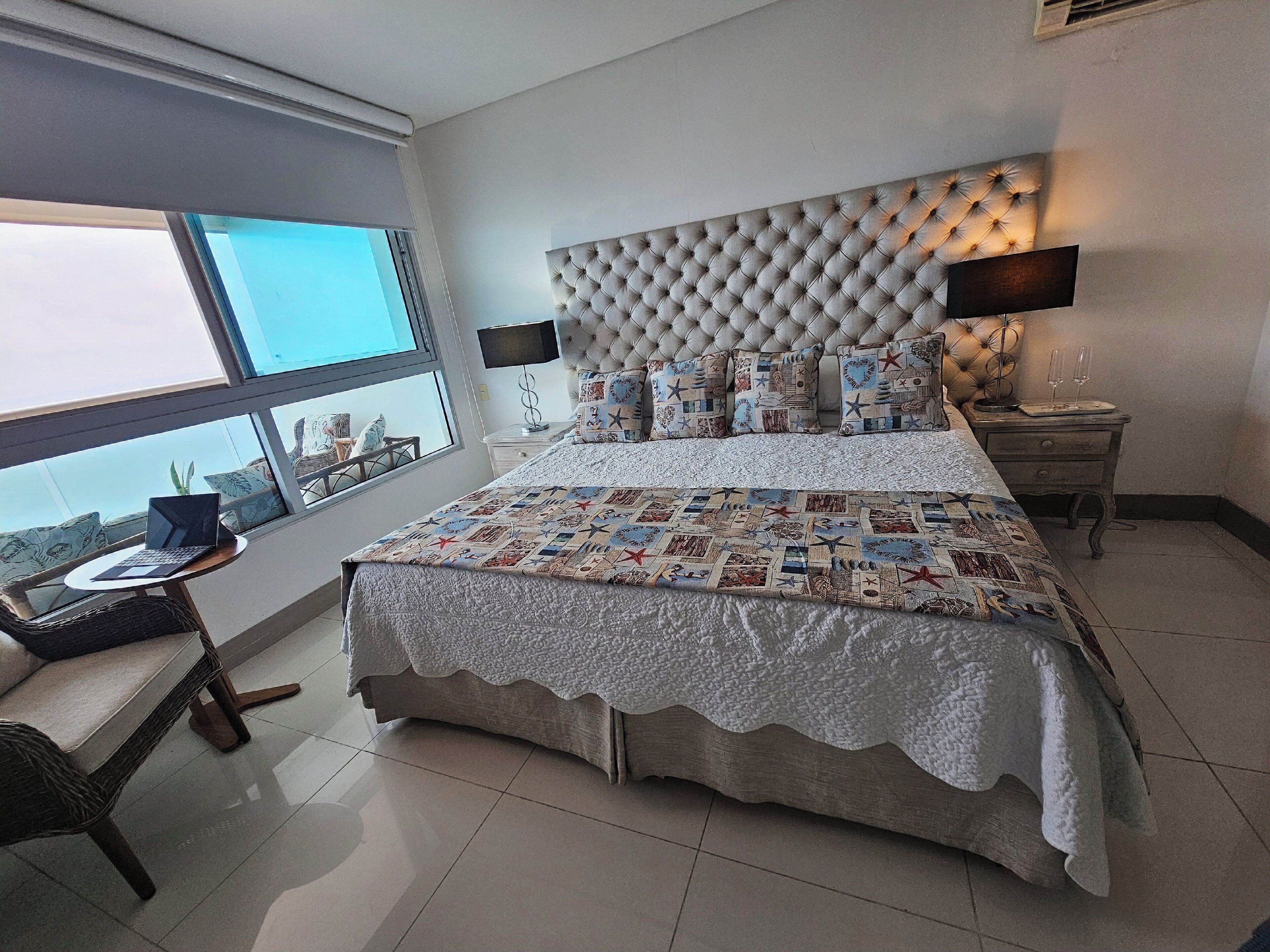 Une chambre moderne avec un grand lit, un bureau avec un ordinateur portable, une fenêtre donnant sur un beau paysage et une table de chevet avec une lampe.